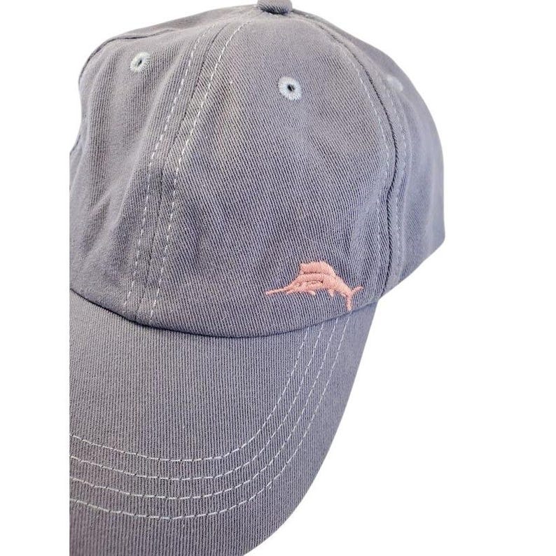 May include: A gray baseball cap with a curved brim and a small, pink embroidered fish logo on the front. The cap has a six-panel design with white stitching and a small button on top. The hat is made of a soft, durable fabric.
