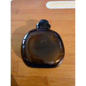 May include: A rounded, dark amber glass bottle with a smooth, reflective surface and a short, narrow neck topped with a small, black, textured screw-on cap. The bottle has a wide, flattened, circular body shape and is made of translucent brown glass.