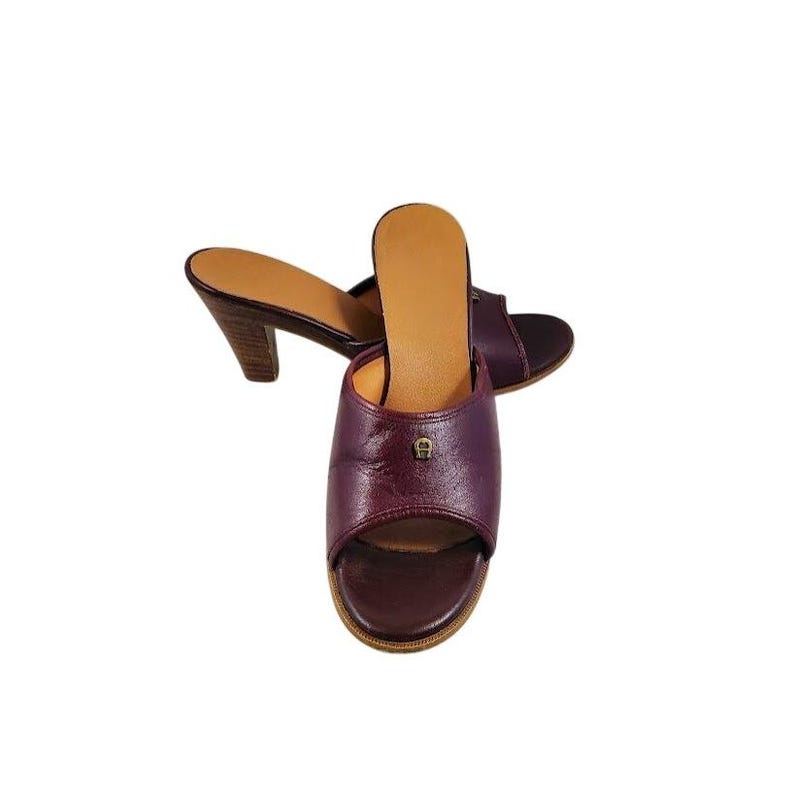 May include: Burgundy leather open-toe slip-on sandals with a small gold emblem. The sandals have a wooden heel and a light brown insole. The shoes are set against a white background.