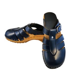 Op de afbeelding: Een paar marineblauwe klompen met houten zolen. De leren bovenstukken hebben een T-bandontwerp met zilverkleurige klinknagels en een gespsluiting. De houten zolen zijn lichtbruin.