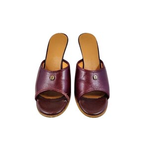 May include: A pair of burgundy leather open-toe slip-on sandals. Each sandal features a small gold-colored emblem on the vamp and a tan insole. The heels are approximately 2 inches high.