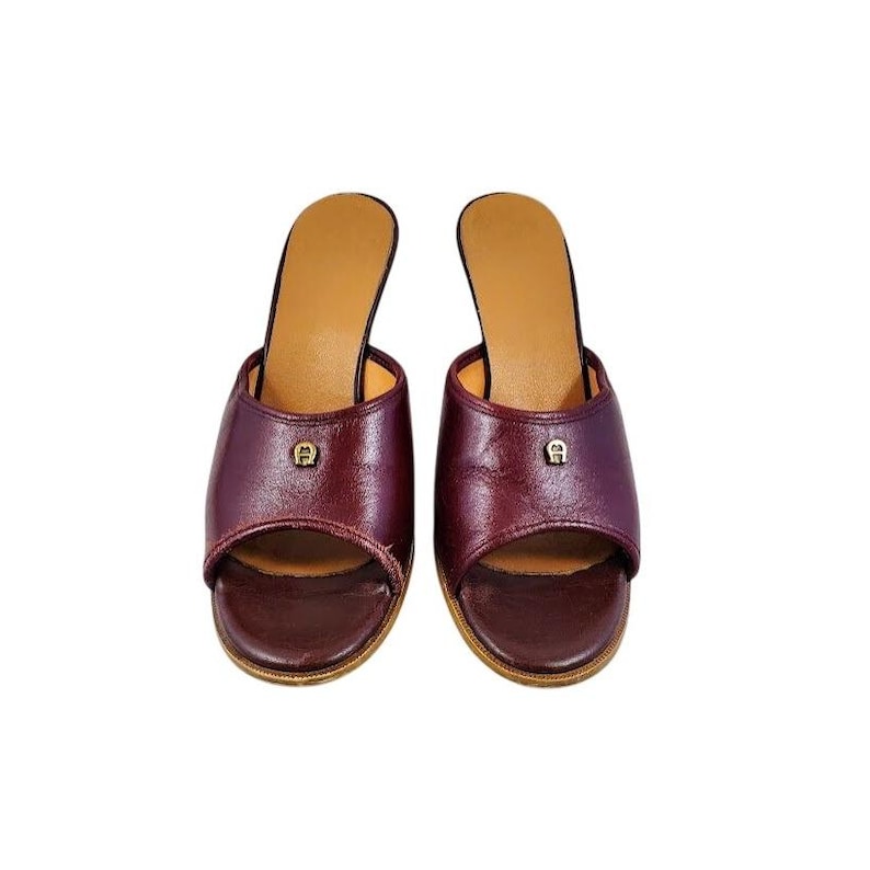 May include: A pair of burgundy leather open-toe slip-on sandals. Each sandal features a small gold-colored emblem on the vamp and a tan insole. The heels are approximately 2 inches high.