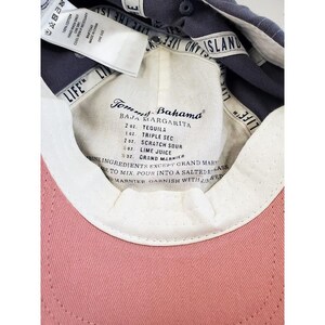 May include: A pink baseball cap with a white interior and a navy blue brim. The interior features a label with a cocktail recipe for a Baja Margarita. The cap is made of 100% cotton.