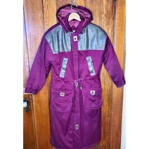 May include: A long, purple winter coat with a hood and silver accents. The coat features a drawstring waist, two front pockets, and a silver panel across the shoulders. The coat is hanging on a wooden door.