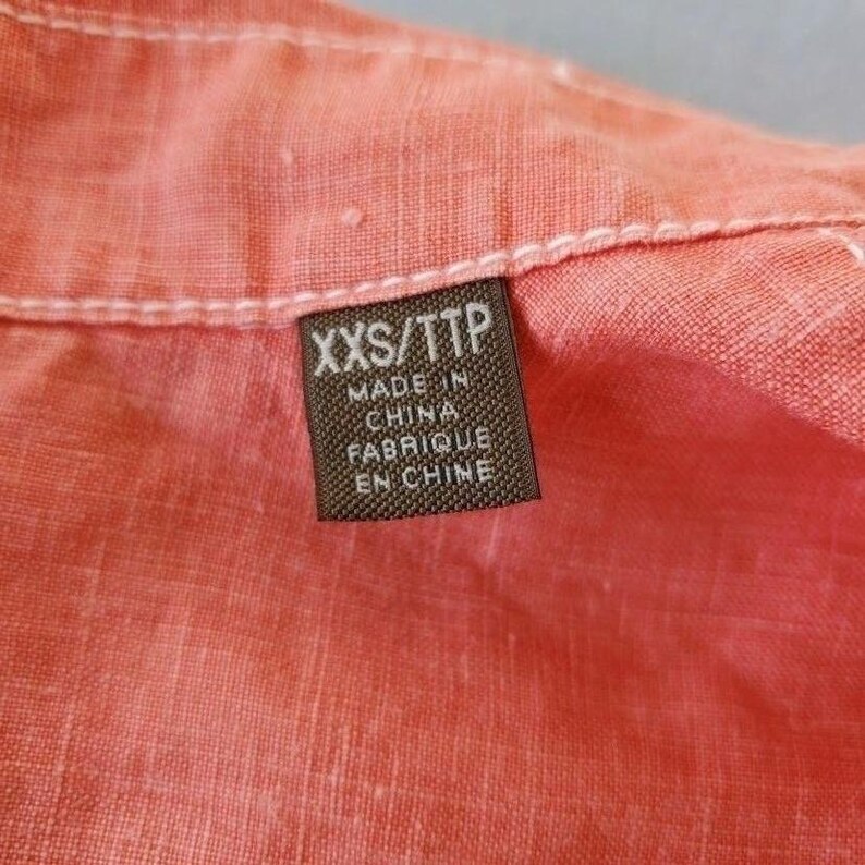 May include: A brown tag with white stitching, attached to a pink fabric garment. The tag reads "XXS/TTP" and "Made in China Fabrique en Chine".