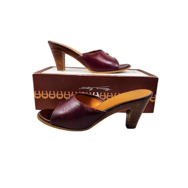 May include: Burgundy leather slip-on mules with wooden heels. The open-toe shoes are displayed on a brown box. The heels are approximately 3 inches high. The shoes have a classic, elegant design.
