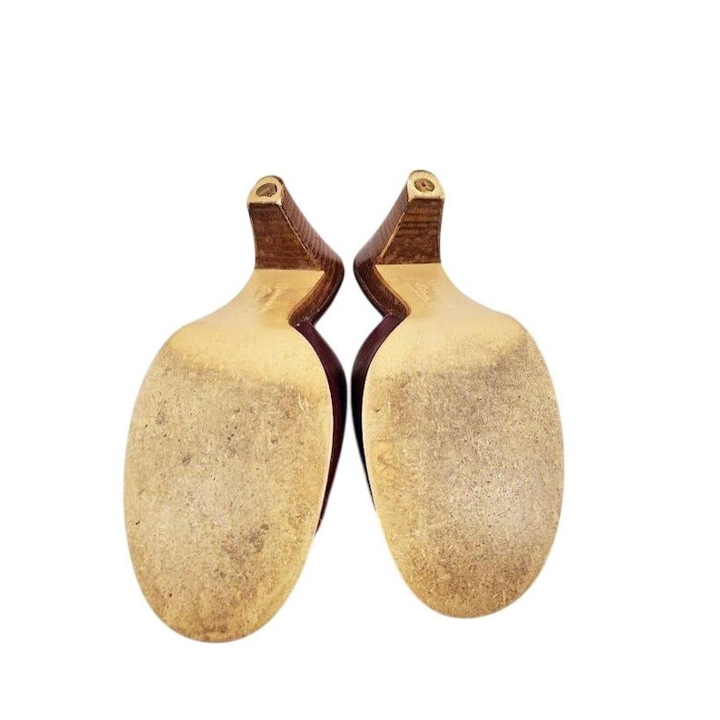 May include: A pair of burgundy-colored high heels, viewed from the soles. The heels are a dark brown color, and the soles are a light tan with wear marks. The shoes are set against a white background.