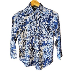 Puede incluir: Camisa abotonada con estampado de cachemir azul y blanco. La camisa de manga larga está hecha de una tela ligera y presenta un cuello clásico y puños abotonados. Cuelga de una percha de madera.