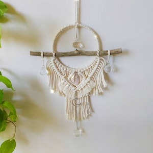 Op de afbeelding: Een witte macramé wandhanger met een houten tak, kristallen en een halve maan bedel.
