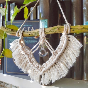 May include: A macrame wall hanging with a crystal in the center, hanging from a branch. The macrame is cream colored and has a fringe around the edges. The crystal is clear and has a diamond shape. The wall hanging is hanging in front of a bookshelf with books.