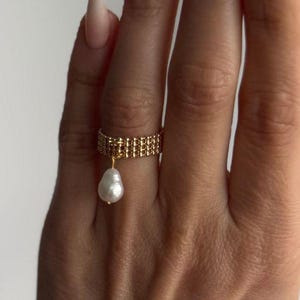 May include: A gold-colored ring with a beaded band and a dangling white pearl. The ring is worn on a finger, showcasing its delicate design and elegant simplicity. The ring's warm hue contrasts with the skin tone, highlighting the pearl's luster.