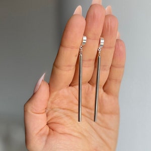 Op de afbeelding: Een paar zilveren oorhangers in de vorm van staven. Elke oorbel heeft een lange, rechte staaf die aan een kleine, cirkelvormige oorbel hangt. De oorbellen worden in een hand gehouden tegen een zachte achtergrond. De tekst "WILYMI JEWELRY CO" staat onderaan.