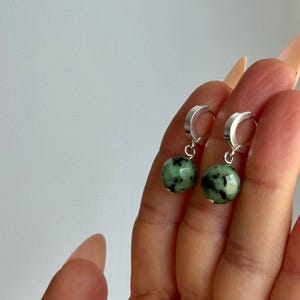 Peut inclure: Boucles d'oreilles créoles en argent avec des perles rondes en pierre verte et noire. Les boucles d'oreilles sont présentées sur un fond clair, mettant en valeur le contraste des matériaux.