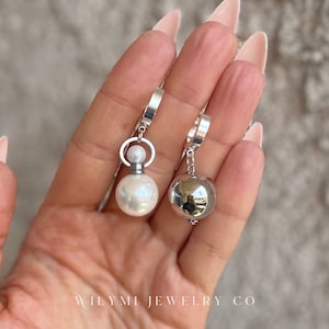 May include: A pair of silver earrings. One earring features a large, white pearl and a smaller pearl, while the other has a reflective silver ball. The earrings are suspended from silver hoops. The text "WILYMI JEWELRY CO" is visible at the bottom.