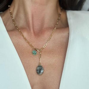 May include: A gold-coloured necklace with a paperclip chain design. The necklace features two pendants: a small, round turquoise stone and a larger, teardrop-shaped clear stone with a gold-coloured bezel. The necklace is worn on a person wearing a white top.