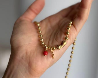 May include: A gold-colored beaded necklace with a small red bead and a charm. The necklace is held in a hand. The text "WILYMI JEWELRY CO" is at the bottom of the image.