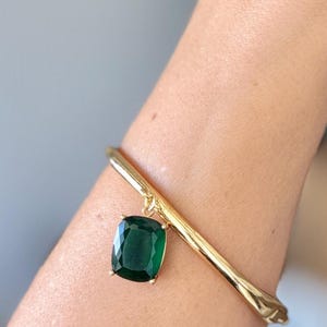 May include: A gold-toned metal bangle bracelet featuring a single, large, cushion-cut dark green gemstone charm suspended from a small circular link. The bracelet has a smooth, polished finish and a hinged clasp mechanism visible on the side.