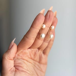 Puede incluir: Dos pendientes tipo ear jacket en tono dorado con un diseño de barra metálica curva. Cada pieza está adornada con dos perlas esféricas blancas, una situada en la parte superior y otra en la inferior del alambre curvo.