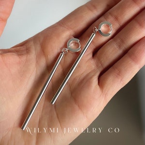 Op de afbeelding: Een paar zilverkleurige oorhangers, elk met een rechte, langwerpige staaf die aan een kleine, cirkelvormige lus hangt. De oorbellen worden getoond tegen een neutrale achtergrond. De tekst "WILYMI JEWELRY CO" is zichtbaar.