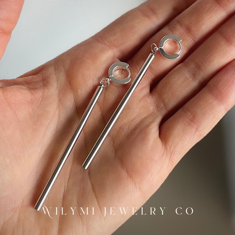 Op de afbeelding: Een paar zilverkleurige oorhangers, elk met een rechte, langwerpige staaf die aan een kleine, cirkelvormige lus hangt. De oorbellen worden getoond tegen een neutrale achtergrond. De tekst "WILYMI JEWELRY CO" is zichtbaar.