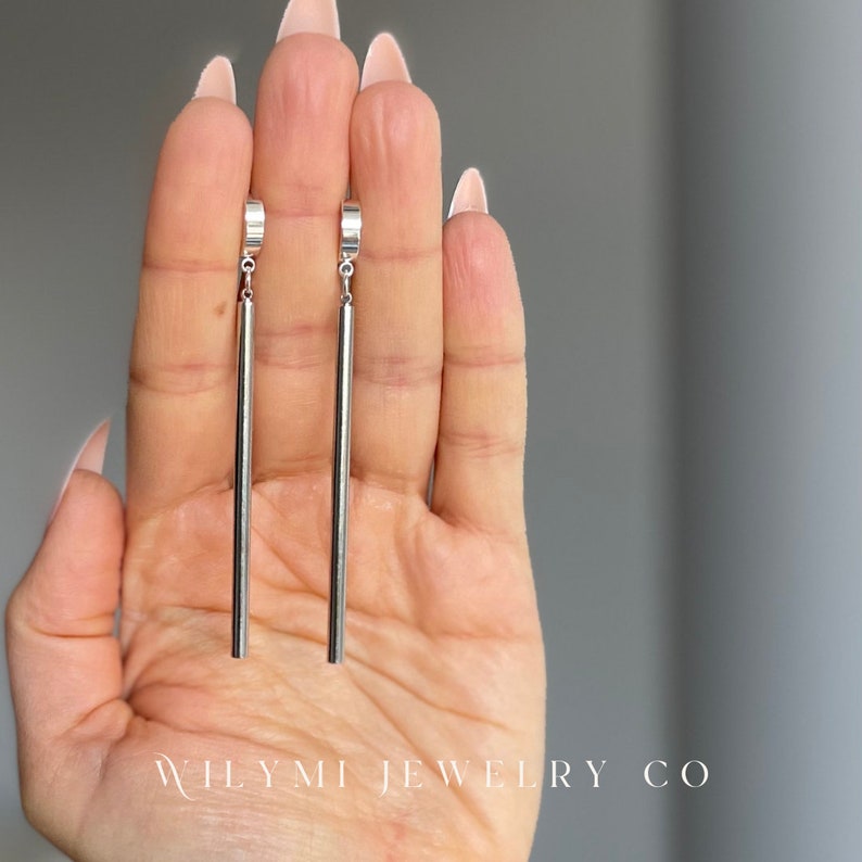 Op de afbeelding: Een paar zilverkleurige oorhangers. Elke oorbel heeft een dunne, rechte staaf die is opgehangen aan een kleine, gebogen bovenkant. De achtergrond is neutraal. De tekst "WILYMI JEWELRY CO" is onderaan zichtbaar.
