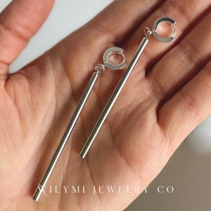Op de afbeelding: Een paar zilverkleurige lineaire oorhangers. Elke oorbel heeft een rechte, langwerpige staaf die is opgehangen aan een kleine, cirkelvormige lus. De oorbellen worden getoond tegen een neutrale achtergrond. De tekst "XILYMI JEWELRY CO" is onderaan zichtbaar.