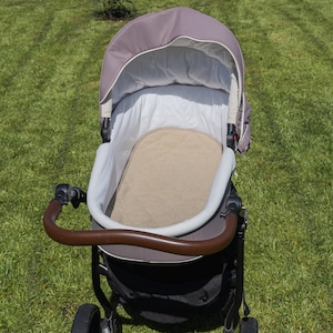 Könnte beinhalten: Ein brauner und schwarzer Kinderwagen mit braunem Griff, beigem Innenraum und braunem Verdeck. Der Kinderwagen steht auf einer grünen, grasbewachsenen Fläche.