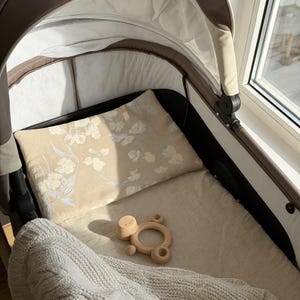 May include: A baby stroller interior featuring a beige floral pillow, a wooden toy, and a cream blanket. The stroller has a brown frame and white lining. The stroller is positioned near a window.