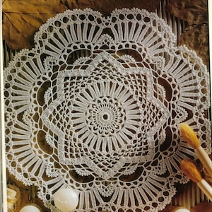 Puede incluir: Un delicado tapete blanco de ganchillo con un diseño circular. El tapete presenta un patrón detallado de calados y elementos en relieve, creando una apariencia delicada y texturizada. El tapete se encuentra sobre una superficie con hojas secas y otros elementos decorativos.