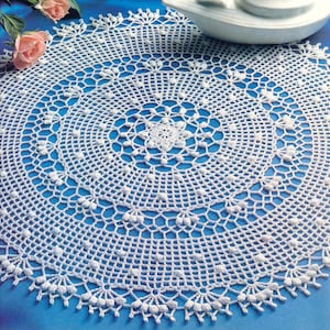 May include: A white, circular, crocheted doily with an intricate openwork design. The doily features a central starburst pattern, surrounded by concentric circles of delicate lace. The edges are finished with a scalloped border. A few pink roses are visible in the background.