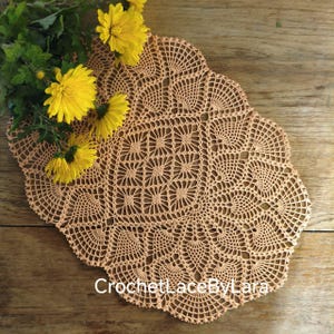 May include: A peach-colored, oval-shaped, crocheted doily with a detailed openwork design. The doily is placed on a wooden surface and is accompanied by a sprig of yellow flowers. The text "CrochetLaceByLara" is visible on the doily.