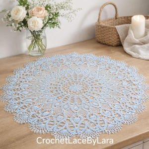 Puede incluir: Un tapete redondo de ganchillo azul claro y blanco con un intrincado diseño floral. El tapete hecho a mano está sobre una superficie de madera. El texto "CrochetLaceByLara" es visible en la parte inferior.