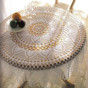 Puede incluir: Mantel de mesa redondo de crochet blanco con un delicado patrón de encaje. El mantel está puesto sobre una mesa redonda de madera clara.