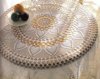 May include: A round wooden table covered with a white crocheted lace tablecloth. The tablecloth features a detailed openwork design with heart-shaped patterns. A plate with fruit and two glasses are on the table.