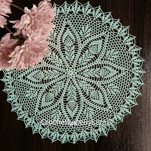 May include: A round, light blue crocheted doily with a detailed floral pattern. The doily is placed on a dark brown wooden surface, with pink flowers in the upper left corner. The doily has a scalloped edge and the text "CrochetLaceByLara" is visible.