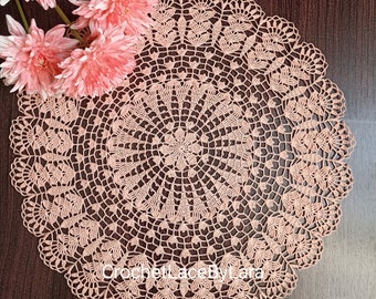 May include: A delicate peach-coloured crochet doily with a circular design. The doily has a centre with a flower-like pattern and intricate lacework around the edges.