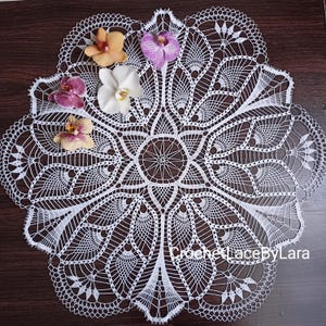 Op de afbeelding: Wit gecrochet doily met een delicaat bloemenpatroon. Het doily is rond en heeft een centraal stervormig patroon. Er zijn vier witte bloemen met roze centra en drie oranje bloemen met gele centra op het doily.