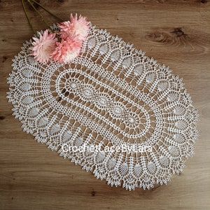 Könnte beinhalten: Ein weißes, oval geformtes, gehäkeltes Deckchen mit aufwendigen Lochmustern. Das Deckchen liegt auf einer Holzoberfläche und ist mit rosa Blumen geschmückt. Der Text "Crochet Lace by Lara" ist auf dem Deckchen zu sehen.