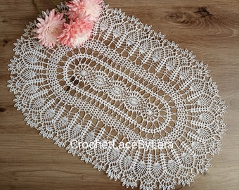 Può includere: Una tovaglietta ovale bianca all'uncinetto con intricati motivi traforati. La tovaglietta è posizionata su una superficie di legno e adornata con fiori rosa. Il testo "Crochet Lace by Lara" è visibile sulla tovaglietta.
