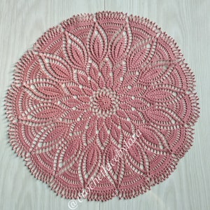 May include: A round, dusty rose crochet doily with a detailed floral design. The doily features a central flower pattern surrounded by intricate leaf-like shapes and a scalloped edge. The text "@crochetlacebylara" is visible.