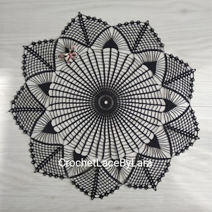 Könnte beinhalten: Schwarze, gehäkelte Tischdecke mit detailliertem, geometrischem Design. Die Tischdecke hat ein zentrales kreisförmiges Muster mit radialen Linien und einem dekorativen Rand. Eine kleine, mit Juwelen besetzte Schmetterlingsnadel liegt auf der Tischdecke. Der Text "CrochetLaceByLara" ist sichtbar.