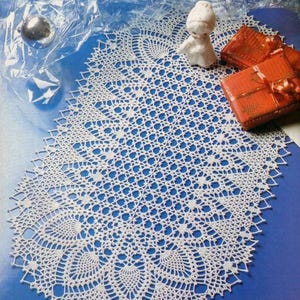 May include: A white, intricately crocheted doily with a scalloped edge, displayed on a blue surface. The doily features a detailed openwork pattern. Decorative elements include a small porcelain figurine and two small, wrapped orange gift boxes.