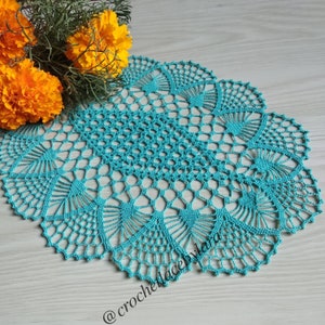May include: A turquoise crochet doily featuring an intricate, repeating fan-like pattern with a central hexagonal mesh design. The delicate, circular lace piece displays a scalloped edge and is marked with the text @crochelacelora.