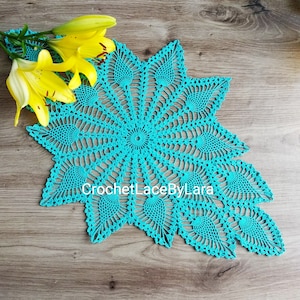 Peut inclure: Nappe en crochet turquoise en forme d'étoile, avec des motifs ajourés complexes. La nappe est posée sur une surface en bois, avec des lys jaunes dans le coin supérieur gauche. Le texte "CrochetLaceByLara" est visible.