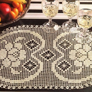 Pode incluir: Um corredor de mesa oval branco de crochê com um delicado padrão floral. O corredor está colocado sobre uma superfície escura com três taças de vidro transparente cheias de um líquido claro.