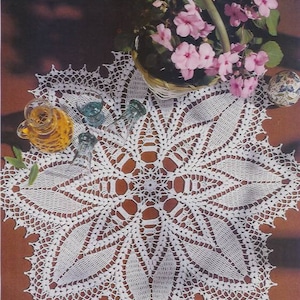 May include: A white, handmade crochet doily with a detailed floral design. The doily is placed on a brown surface and is surrounded by a basket of pink flowers, small glasses, and a decorative glass bottle.