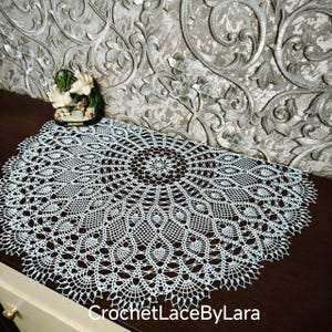 May include: A light blue, intricately crocheted doily with a detailed floral pattern. The doily is circular and rests on a dark brown surface. The text "CrochetLaceByLara" is visible at the bottom right.