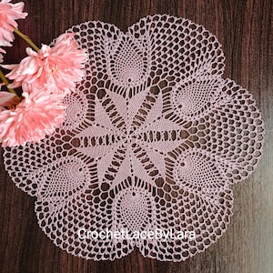 May include: A detailed, light pink crocheted doily with a floral design. The doily features a central starburst pattern surrounded by circular motifs and a scalloped edge. The text "CrochetLaceByLara" is visible. Pink flowers are in the upper left corner.