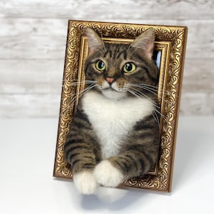 Un gato atigrado marrón hecho a mano con lana fieltrada con el pecho y las patas blancas, colocado dentro de un marco dorado rectangular y ornamentado. El gato tiene ojos verdes, largos bigotes blancos y nariz rosa, pareciendo asomarse fuera del marco.