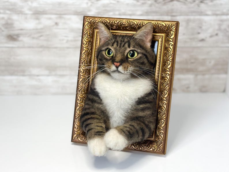 Un gato atigrado marrón hecho a mano con lana fieltrada con el pecho y las patas blancas, colocado dentro de un marco dorado rectangular y ornamentado. El gato tiene ojos verdes, largos bigotes blancos y nariz rosa, pareciendo asomarse fuera del marco.